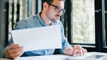 Serious pensive thoughtful focused young casual business accountant bookkeeper in office looking at and working with laptop and income tax return papers and documents